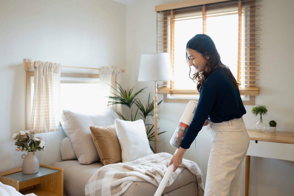 Woman vacuuming a sofa with a cordless handheld vacuum in a bright living room, illustrating turnover cleaning